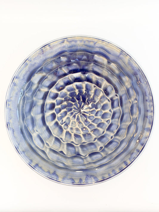 A round ceramic plate with a textured, concentric ripple pattern in blue and white glaze, photographed from directly above on a white background.